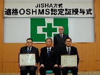 OSHMS認定証を持つ富岡聖新城副工場長（左端）と高塚義美安全衛生課係長（右端）。後列は安全衛生課の今泉紀夫。写真中央は中央労働災害防止協会の関澤秀哲理事長。